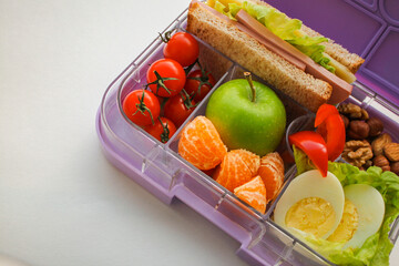 Lilac lunch box with compartments in which-useful food for lunch and snack: sandwich, vegetables, fruits, nuts on a white background. Concept of healthy food, snack for adults and children