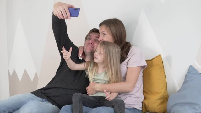 Happy Russian family with small cute kid daughter sit on sofa make photos with smart phone.