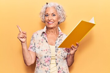 Senior grey-haired woman holding book smiling happy pointing with hand and finger to the side