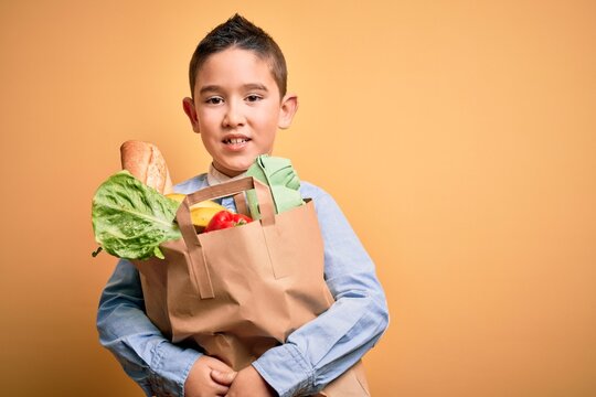 Adorable toddler holding paper bag with food standing over isolated yellow background