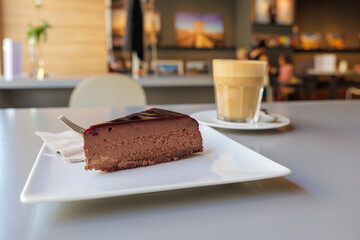 Selected focus view at simply chocolate cheese cake on white plate beside with a cup of coffee, on table beside window of bright cafe.