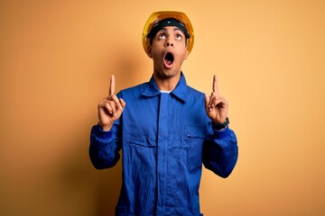 Young handsome african american worker man wearing blue uniform and security helmet amazed and surprised looking up and pointing with fingers and raised arms.