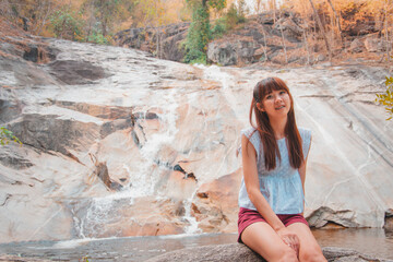 Asian woman travelers are smiling and relaxes sitting on the stone in front of the waterfall at Namtok Kaew Chan., Ratchaburi, Thailand.