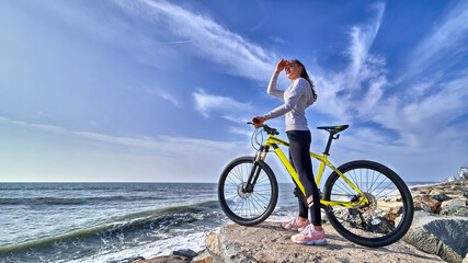 Fitness woman with a bicycle stands on the stones on the seashore and looks into the distance on a...