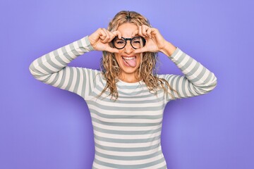 Beautiful blonde woman wearing casual striped t-shirt and glasses over purple background doing ok gesture like binoculars sticking tongue out, eyes looking through fingers. Crazy expression.