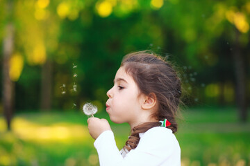 Portrait of prettyl little Girl blowing dandelion flower and smiling in summer park. Happy cute child having fun outdoors.