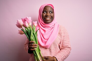Young african american plus size woman wearing muslim hijab holding bouquet of pink tulips smiling looking to the side and staring away thinking.