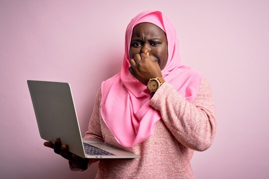 African american plus size woman wearing muslim hijab using laptop over pink background smelling something stinky and disgusting, intolerable smell, holding breath with fingers on nose. Bad smell