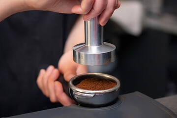 close-up photo of female hands holding a metal tamper and a portafilter with coffee in a coffee shop. A barista preparing for pressing ground coffee for brewing espresso or americano in a cafe