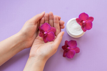 Cosmetic background with hands, soft white cream and fragile magenta colored orchids.Rejuvenating facial cream in a jar and pink orchid flowers on purple background, skin care cosmetics.