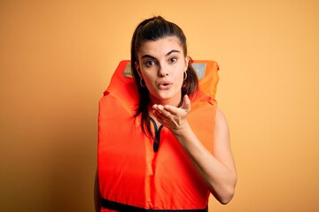 Young beautiful brunette woman wearing orange safe lifejacket over yellow background looking at the camera blowing a kiss with hand on air being lovely and sexy. Love expression.