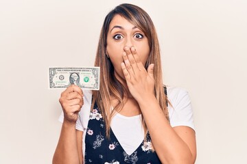 Young beautiful woman holding one dollar covering mouth with hand, shocked and afraid for mistake....