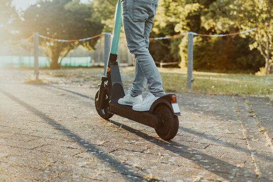 Person f&auml;hrt entlang auf der Stra&szlig;e im Park einen E-Scooter bei Sonnenuntergang. Junger Mensch f&auml;hrt einen elektrischen Cityroller im Sommer mit Hilfe neuster Technologie.