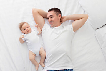 Happily family: father with his son a boy lie on their backs on a white bed in a sunny bedroom. Parent and little child are resting at home. View from above