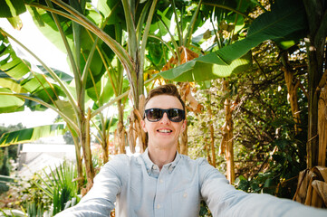 A young man makes a Selfie photo on the background of banana palm trees. In a botanical garden, on the nature in sun rays. sunlight. Close up.