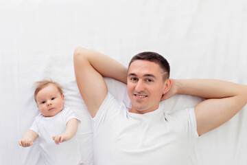 Happily family: father with his son a boy lie on their backs on a white bed in a sunny bedroom. Parent and little child are resting at home. View from above