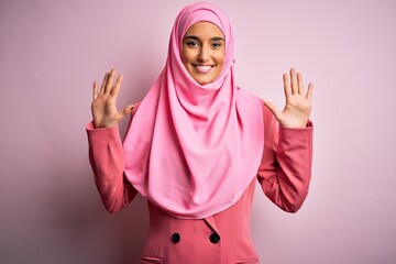 Young beautiful brunette businesswoman wearing pink muslim hijab and business jacket showing and pointing up with fingers number ten while smiling confident and happy.
