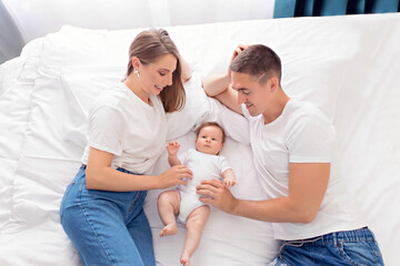 Happy family: father, mother and baby son play on a white bed in a sunny bedroom. Family having fun together, hugging each other.