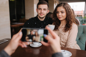 Hand of female making photo of beutiful young couple in cafe. Happy smiled couple posing for photo on smartphone