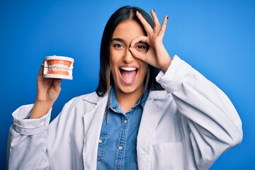 Young beautiful brunette dentist woman wearing coat holding plastic teeth prosthesis with happy face smiling doing ok sign with hand on eye looking through fingers