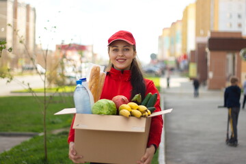 Fototapeta premium Courier girl with a box of food on the street. The girl from the delivery service holds a box in which are: vegetables, fruits, bread, water, milk. She holds the box forward and smiles.