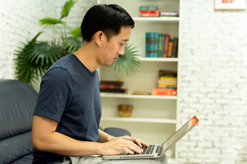 Young smiling Asian man in casual clothing on sofa working on laptop computer. Male freelancer working from home on notebook. Work from home concept