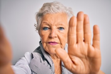 Senior beautiful grey-haired woman making selfie by camera over isolated white background with open hand doing stop sign with serious and confident expression, defense gesture