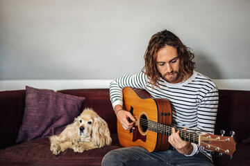 attractive man with long hair with eyes dressed in jeans and in a blue and white striped t-shirt sitting next to a dog of breed american spaniel cocker on a sofa playing acoustic guitar