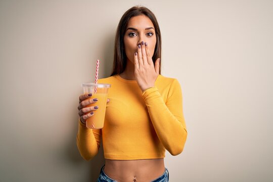 Young beautiful brunette woman drinking healthy orange juice over white background cover mouth with hand shocked with shame for mistake, expression of fear, scared in silence, secret concept
