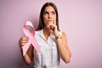 Young beautiful brunette woman holding cancer ribbon symbol over pink background serious face thinking about question, very confused idea