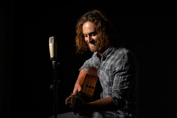 Obraz premium attractive man with long hair with blue eyes sitting next to a microphone and playing acoustic guitar dressed in lumberjack plaid shirt on black background