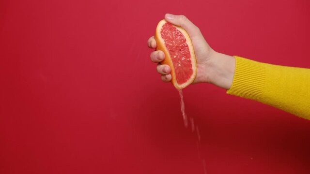 Close up of female hand squeezing fresh sliced grapefruit. Citrus juice drains from pulp and drips, isolated on red studio background with copy space. 