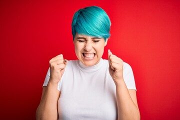 Young beautiful woman with blue fashion hair wearing casual t-shirt over red background excited for success with arms raised and eyes closed celebrating victory smiling. Winner concept.