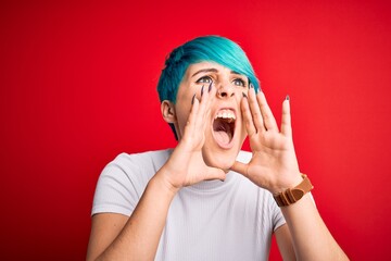 Young beautiful woman with blue fashion hair wearing casual t-shirt over red background Shouting angry out loud with hands over mouth