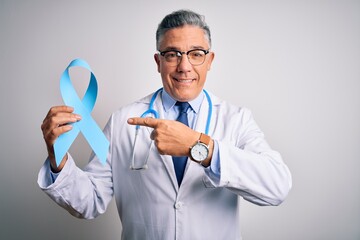 Middle age handsome grey-haired doctor man wearing coat holding blue cancer ribbon very happy pointing with hand and finger