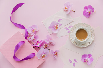 Delicate flatlay composition with morning cup of coffee with milk or cappuccino, letters, pink gift bag and orchid flowers on light pink background.
