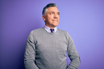 Middle age handsome grey-haired man wearing elegant sweater over purple background looking away to side with smile on face, natural expression. Laughing confident.
