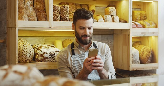 Portrait of happy baker working in bakery shop using smartphone. - Powered by Adobe