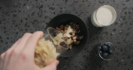 top view man hands preparing breakfast with chocolate granola, almond flakes and blueberry in black bowl on terrazzo surface