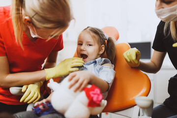 Cheerful little patient with opened mouth looking at camera while female dentist checking teeth near assistant