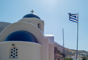 Angle of a traditional Greek orthodox church in blue and white color with a cross at the top and the Greek flag waving on the right