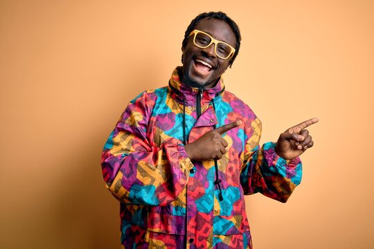 Handsome african american man wearing colorful coat and glasses over yellow background smiling and looking at the camera pointing with two hands and fingers to the side.