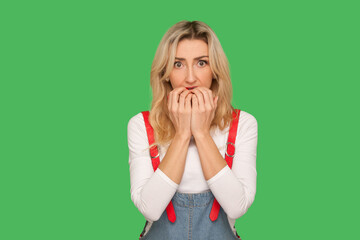 Portrait of nervous stressed adult woman in denim overalls biting her nails and looking with big scared eyes, feeling anxious unsure, having phobia. indoor studio shot isolated on green background