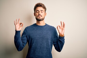 Young handsome man with beard wearing casual sweater standing over white background relax and smiling with eyes closed doing meditation gesture with fingers. Yoga concept.