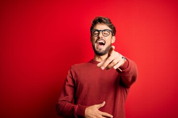 Young handsome man with beard wearing glasses and sweater standing over red background laughing at you, pointing finger to the camera with hand over body, shame expression