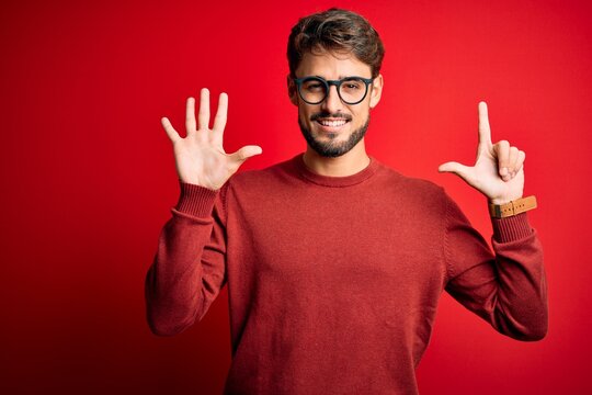 Young handsome man with beard wearing glasses and sweater standing over red background showing and pointing up with fingers number seven while smiling confident and happy.