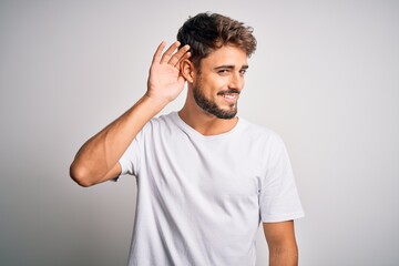 Fototapeta na wymiar Young handsome man with beard wearing casual t-shirt standing over white background smiling with hand over ear listening an hearing to rumor or gossip. Deafness concept.