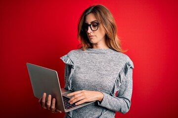 Naklejka premium Young beautiful brunette businesswoman wearing glasses using laptop over red background with a confident expression on smart face thinking serious