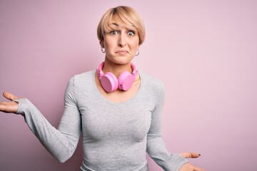 Young blonde woman with short hair wearing headphones on neck over pink background clueless and...