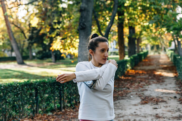 Young athlete woman stretches her left arm on a path in a leafy park